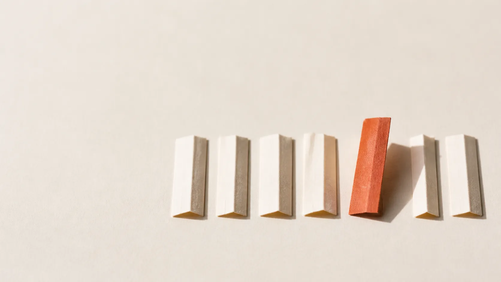 A row of folded paper strips, one lifted and creased in terracotta, singled out from the rest.