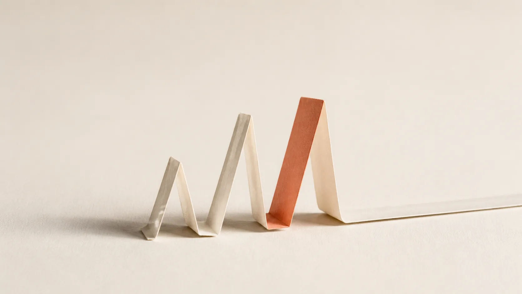 Folded paper climbing in three accordion peaks, the tallest in terracotta, then collapsing flat into a long tail.